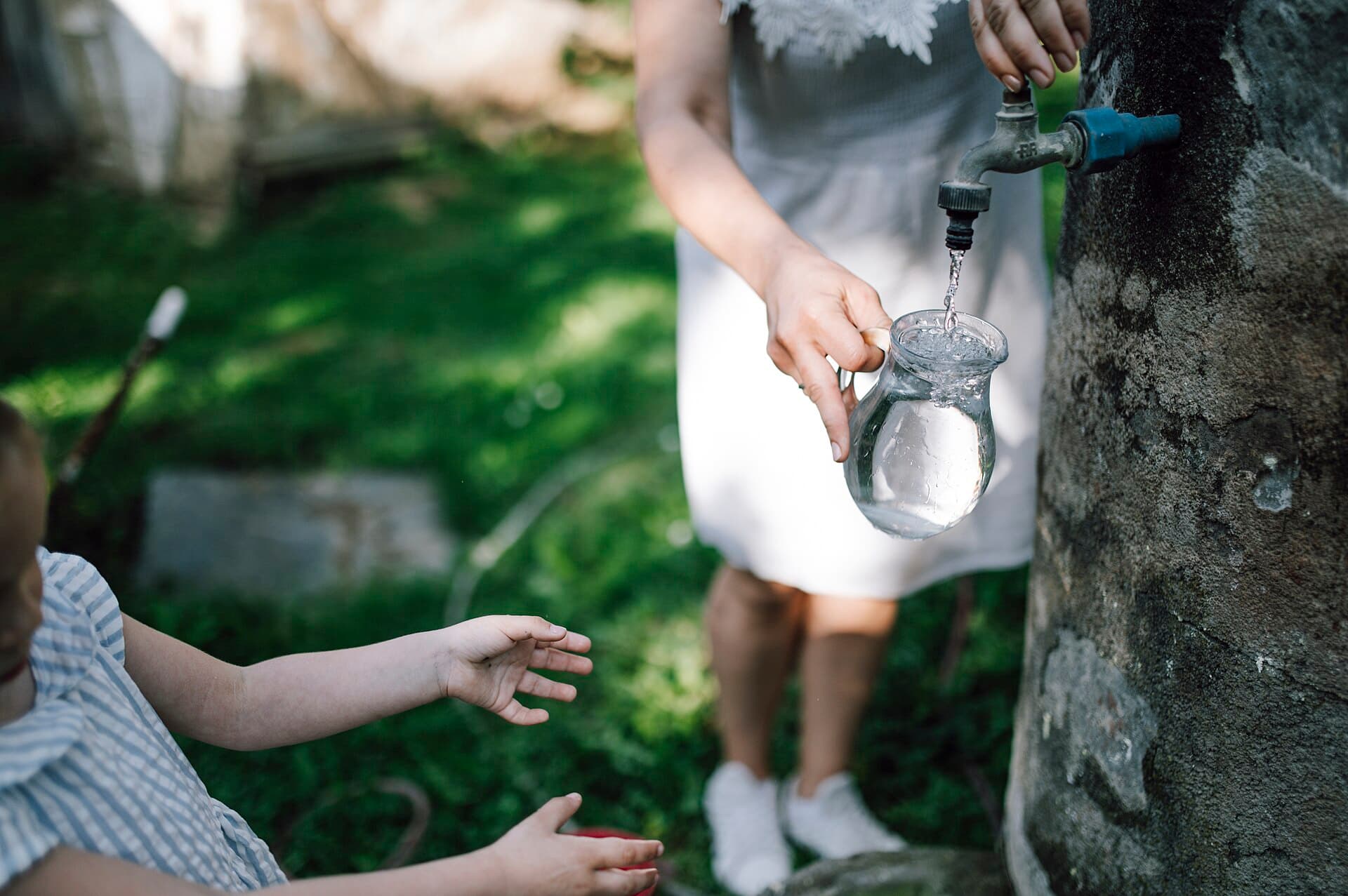 A child reaches for water while an adult fills a glass jar from the tap